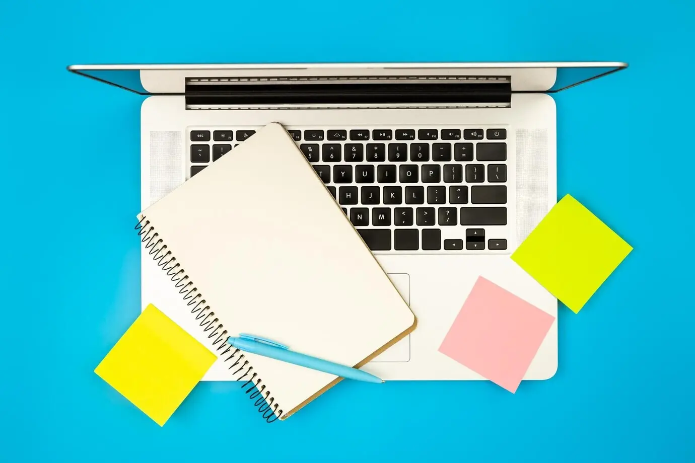 Laptop, Notizblock und Erinnerungszettel aus Papier auf blauem Hintergrund in Draufsicht.